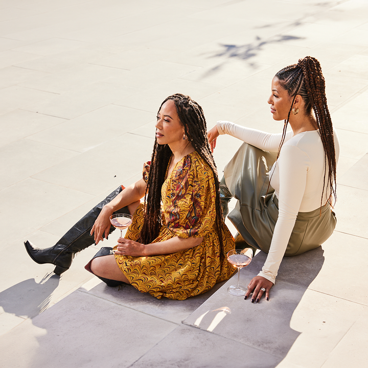 Two women sitting on stone steps outdoors, one wearing a mustard yellow patterned dress and black boots while holding a wine glass, and the other wearing a cream long-sleeve top and olive green skirt.