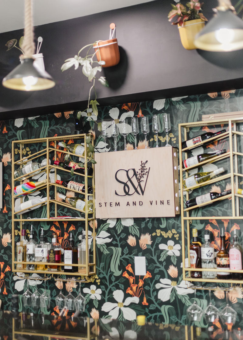 A bar or lounge interior with a dark floral wallpaper backdrop, gold-framed shelving filled with bottles and glassware, pendant lights, and a central sign reading 'Stem and Vine' with a stylized SV logo.