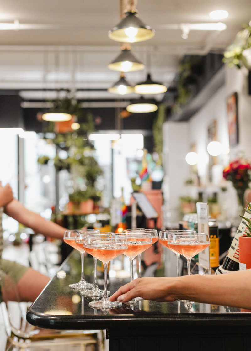 A row of stemmed cocktail glasses filled with pink-orange cocktails on a bar counter, with a bartender's hand resting near the glasses in a softly blurred bar interior.