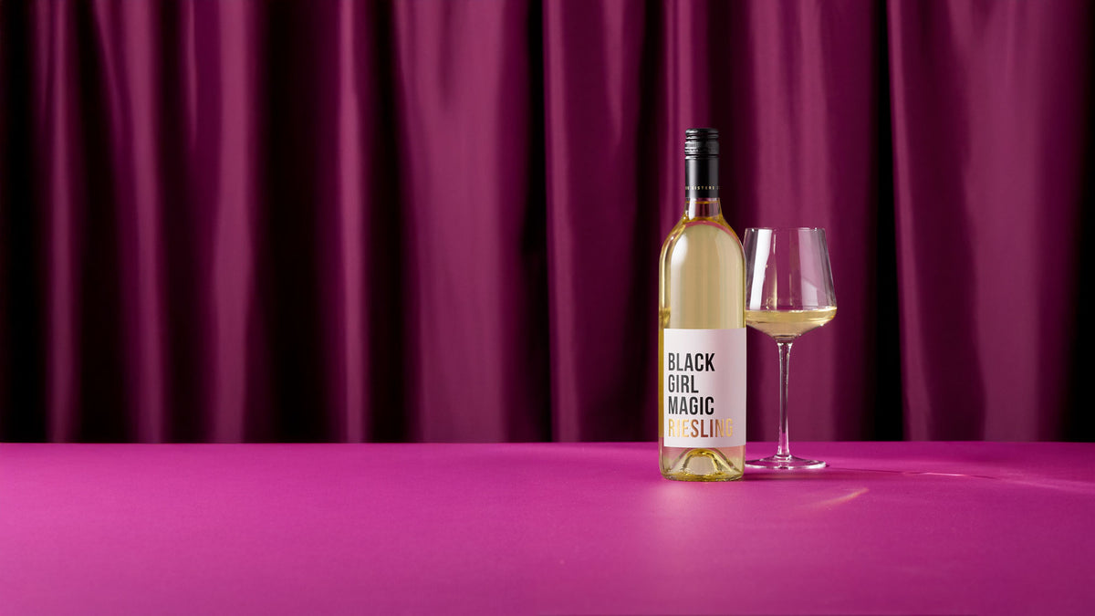 A bottle of Black Girl Magic Riesling wine and a filled wine glass on a bright pink table in front of a draped magenta curtain backdrop.