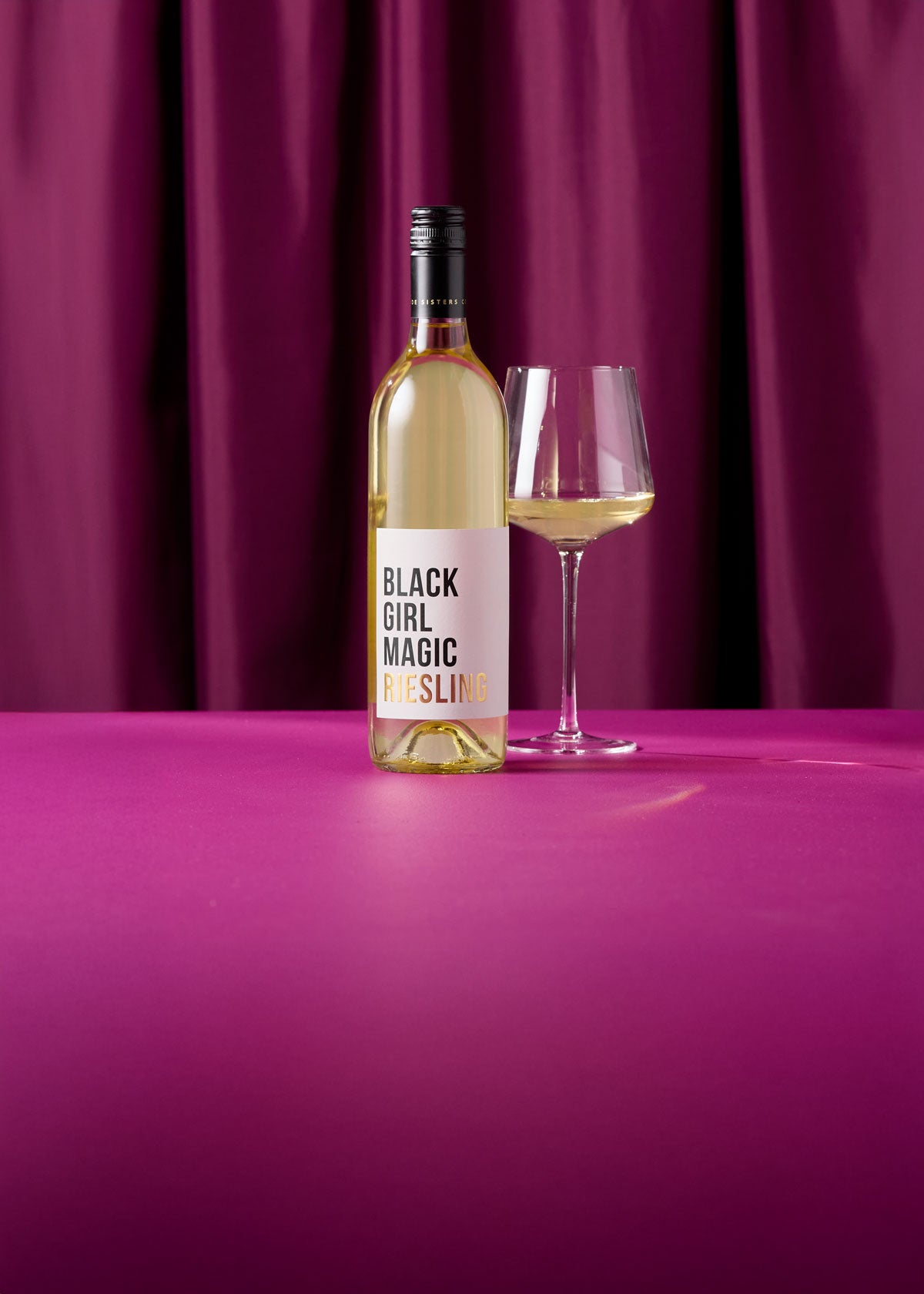A bottle of Black Girl Magic Riesling wine and a filled wine glass on a bright magenta surface in front of a matching purple curtain backdrop.