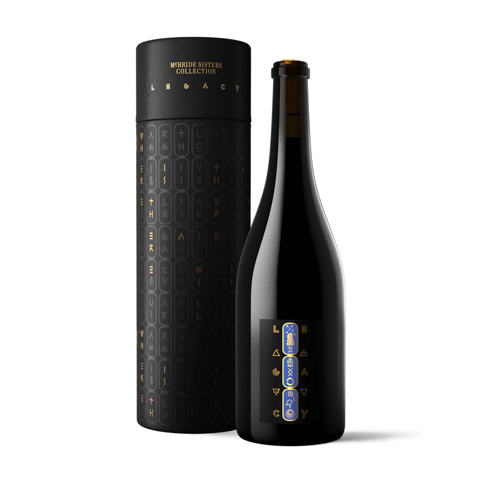 A dark glass bottle with a black label stands beside a tall cylindrical black gift box. Both pieces feature gold lettering and a vertical blue-and-gold monogram design on the bottle label and box lid.