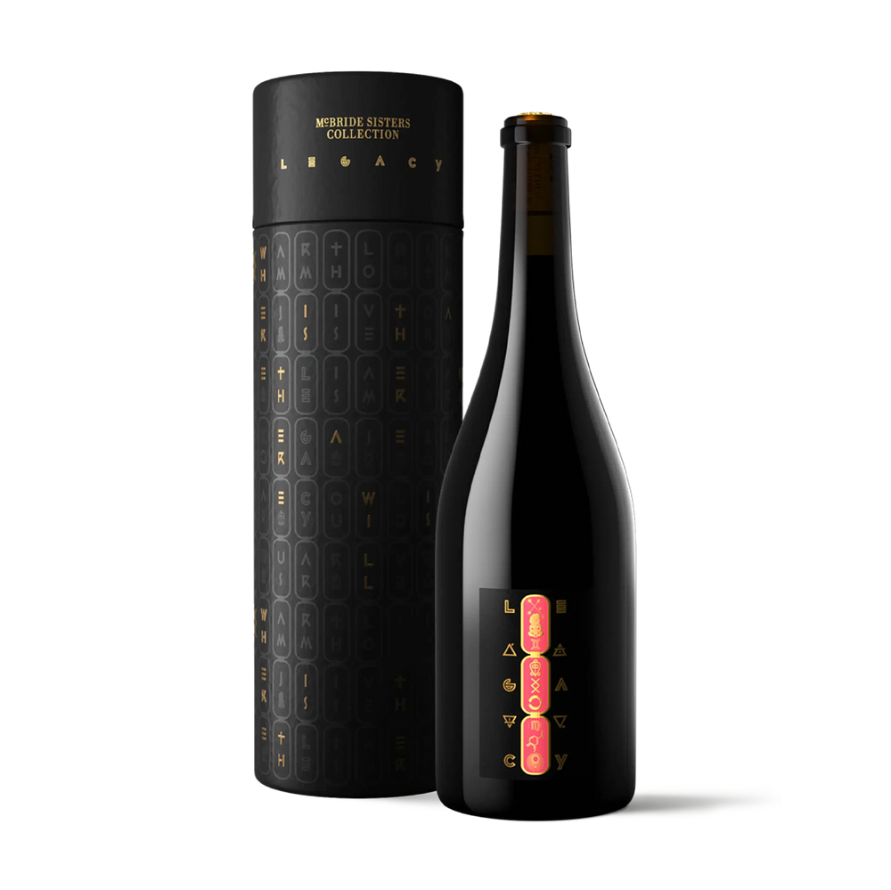 A dark glass bottle with a black label featuring gold and pink lettering stands next to its matching black cylindrical packaging tube, both from the Minnie Sisters Collection Legacy line, on a solid white background.