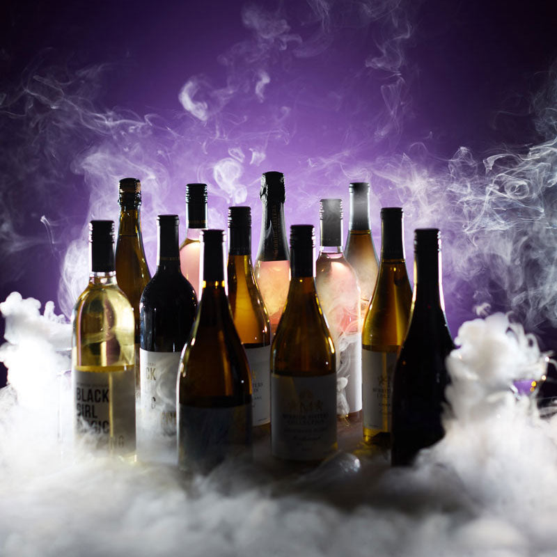 A group of wine bottles arranged in a cluster on a bed of white dry ice, with dramatic purple and blue lighting and smoke in the background.