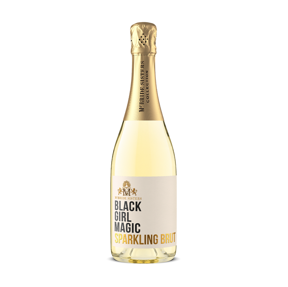 A bottle of McBride Sisters Collection Black Girl Magic Sparkling Brut non-alcoholic sparkling wine, featuring a pale yellow liquid, a gold foil neck, and a cream-colored label with black and gold text, set against a plain white background.