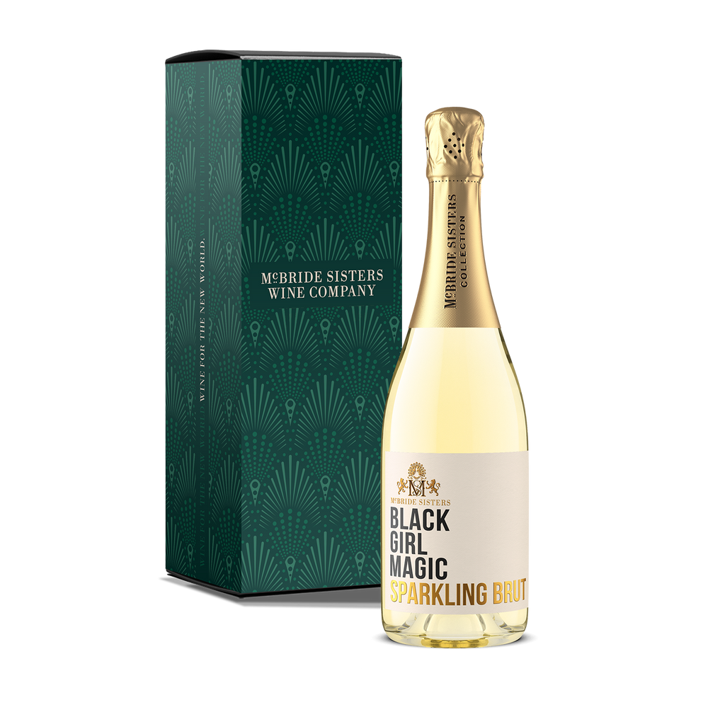A bottle of McBride Sisters Wine Company Black Girl Magic Sparkling Brut next to its dark green gift box with a geometric pattern, on a solid white background.