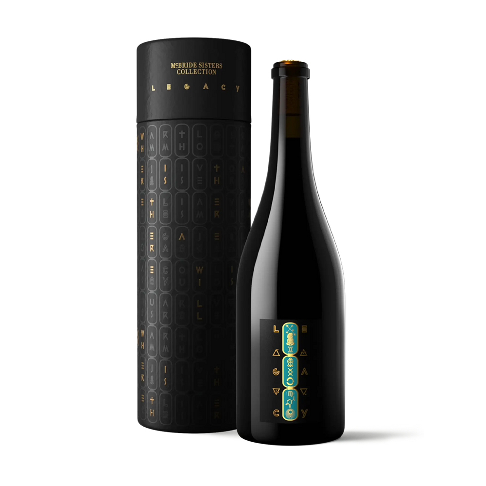 A dark glass bottle with a black label stands beside a tall cylindrical black presentation tube. Both pieces feature gold lettering and a vertical teal and gold graphic near the bottom.
